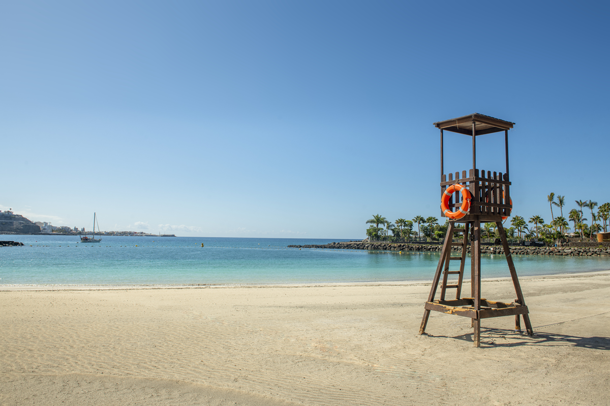 Rettungsturm mit Rettungsring auf hellem Sandstrand, im Hintergrund Palmen, Steinmole und türkisblaues Meer mit Segelboot.
